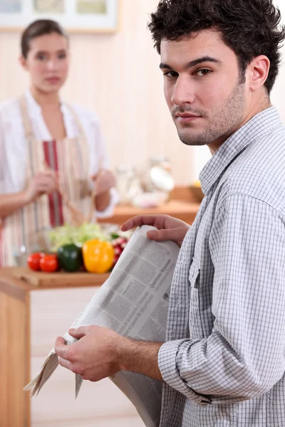 Ung kvinna matlagning och pojkvän läsa tidningen — Stockfoto