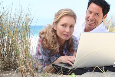 Wife working on laptop on the beach. clipart