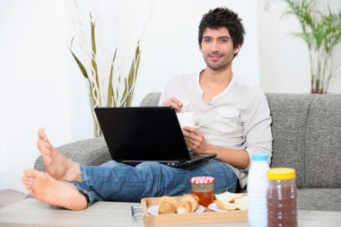 Young man seated on a sofa with legs stretched out, doing computer and eati clipart