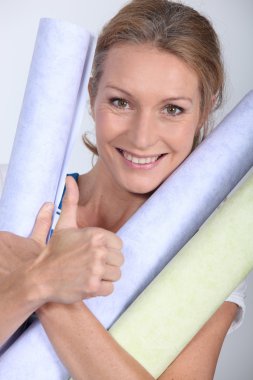 Blond woman carrying rolls of different coloured wallpaper clipart