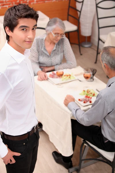 An old couple eating at restaurant and a young waiter — Stock Photo, Image