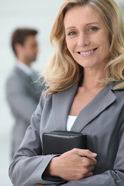 Smiling businesswoman holding personal organiser with colleague in backgrou — Stock Photo, Image