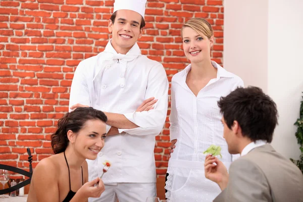 Una pareja cenando en el restaurante, un cocinero y una camarera detrás de ellos — Foto de Stock