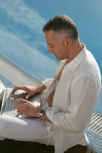 Medelålders gentleman sitter på poolen kanten med laptop — Stockfoto