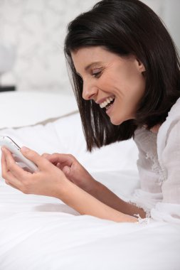 Woman on bed laughing and watching a mobile phone clipart