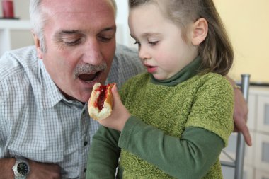 Granddaughter giving her grandfather a piece of bread with jam to eat clipart