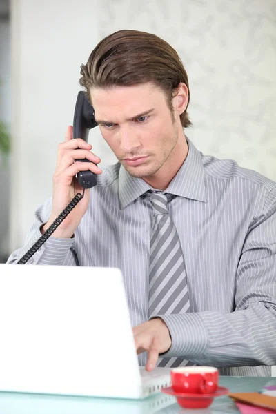 Молодой бизнесмен разговаривает по телефону, пользуясь ноутбуком — стоковое фото