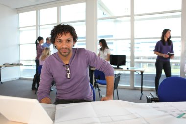 Young man in architect's office with laptop and plans clipart