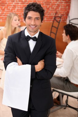 Well dressed waiter facing the camera, behind a couple is dining at restaur clipart