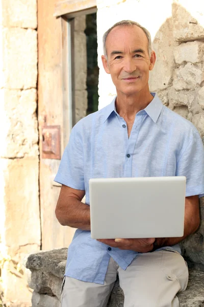 头发灰白的男人拿着笔记本电脑坐在石墙上 — 图库照片
