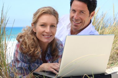 Wife working on laptop on the beach. clipart