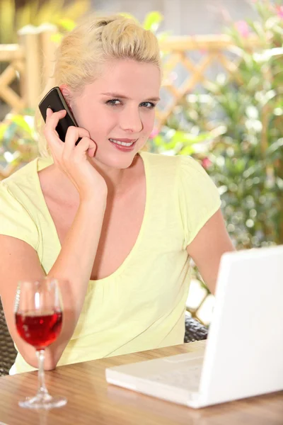 Vrouw praten over haar mobiele telefoon vóór haar laptop en een glas van win — Stockfoto
