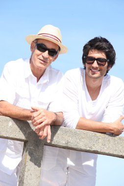 Two men in white leaning on a fence with a blue sky background clipart
