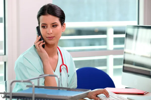 Giovane infermiera al telefono dietro il suo computer — Foto Stock