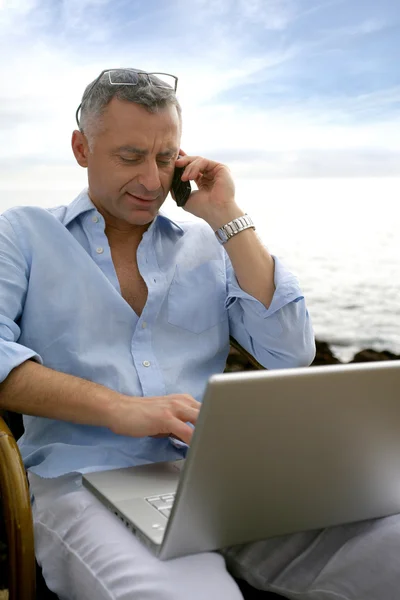 Empresário de férias trabalhando em seu laptop e falando em seu celular — Fotografia de Stock