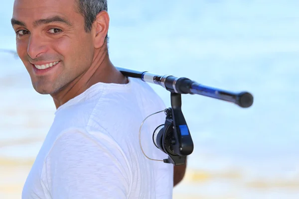 Un hombre de 40 años caminando por la playa con una caña de pescar en el hombro —  Fotos de Stock
