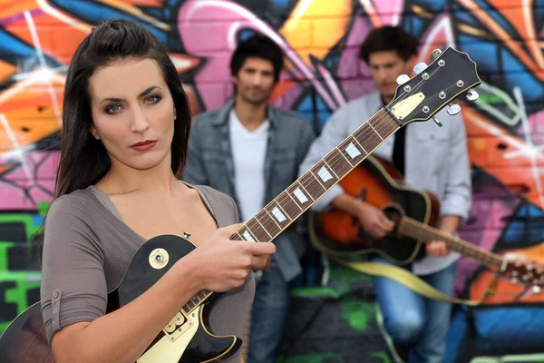 Female guitarist standing with band members in front of a graffitied wall — Stock Photo, Image
