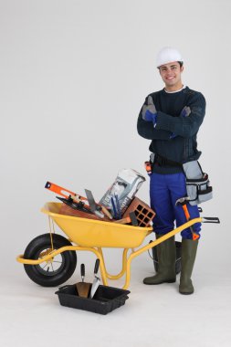 Builder stood by wheelbarrow full of equipment clipart