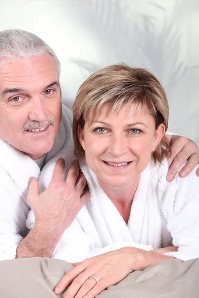Mature couple in bathrobe laid on a bed — Stock Photo, Image
