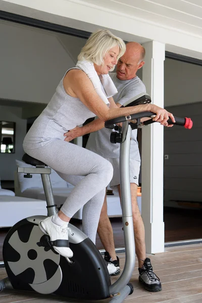 Middle-aged blonde woman on exercise bike coached by husband — Stock Photo, Image