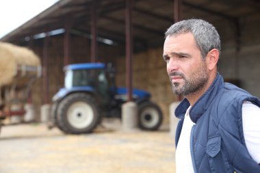 Farmer standing in front of a barn containing a tractor clipart