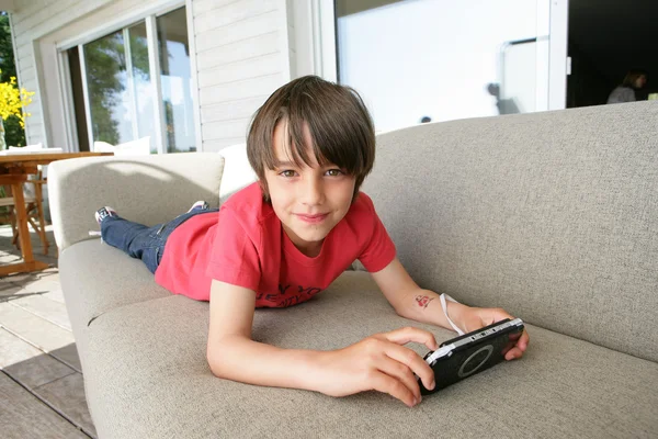 Мальчик лежит на диване с ручной видеоигрой — стоковое фото