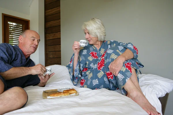 Casal sênior em roupão sentado em uma cama tomando café da manhã — Fotografia de Stock
