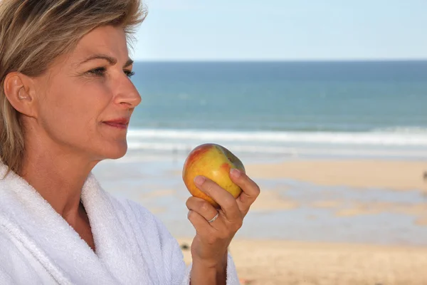 Reife blonde Frau im Bademantel isst einen Apfel vor dem Meer — Stockfoto