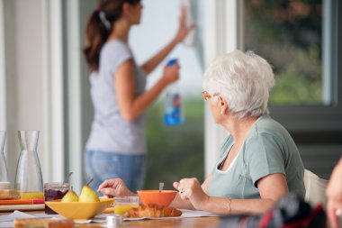 Senior woman having breakfast with home care in the background
