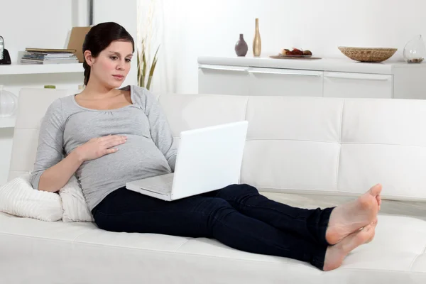 Una donna incinta sul suo divano con un computer portatile . — Foto Stock