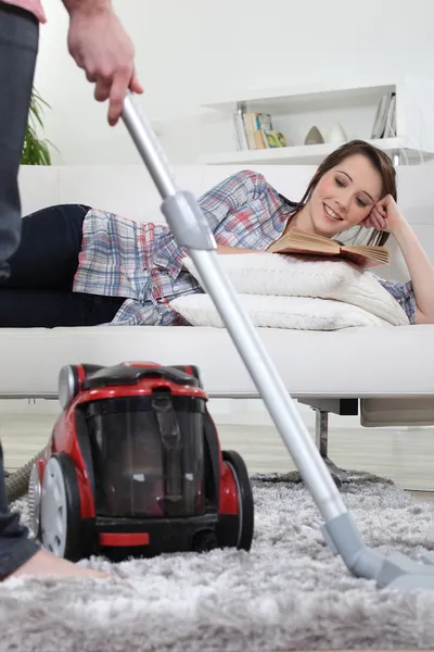 A man vacuuming while his girlfriend is reading a book. — Stock Photo, Image