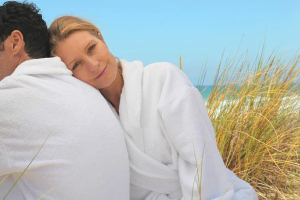 Couple sitting in the sand dunes in white toweling robes — Stock Photo, Image