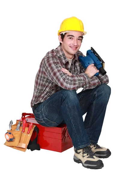 Молодой торговец сидит на ящике с инструментами и держит шлифовальную машину — стоковое фото
