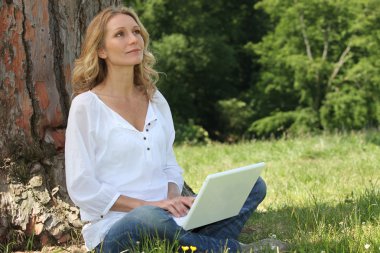 Blond woman sat by tree with laptop deep in thought clipart