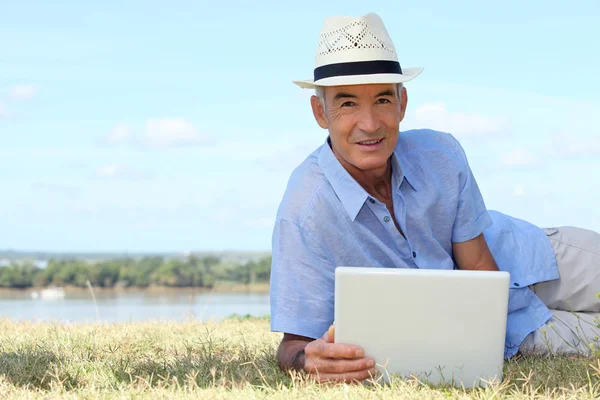 Homem sênior usando um computador portátil em uma margem do rio — Fotografia de Stock