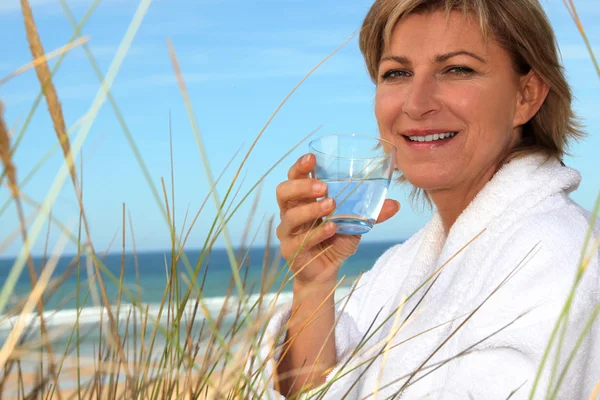 Vrouw met een badjas en een glas water op het strand — Stockfoto