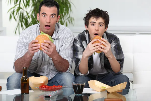 Far och son satt i soffan, äta hamburgare — Stockfoto