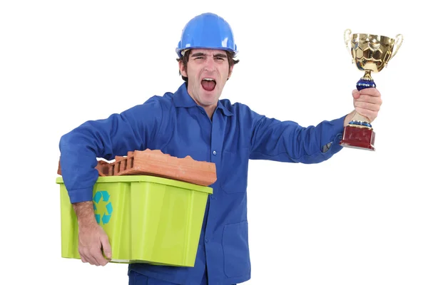 Een bouwvakker holding een trofee en een recycling mand. — Stockfoto