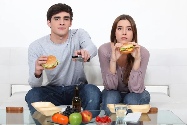 Retrato de jovem casal comendo hambúrgueres enquanto assiste TV — Fotografia de Stock