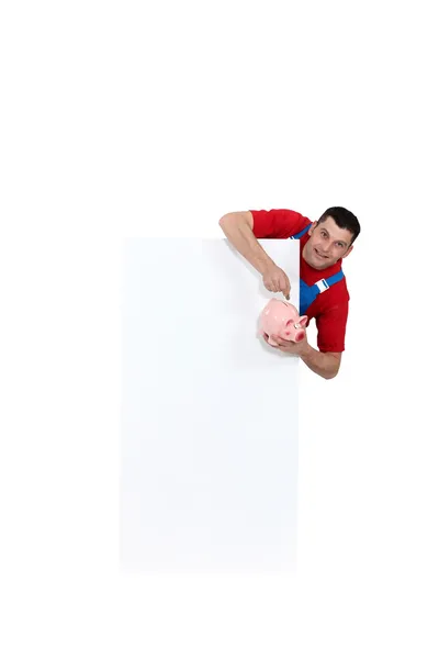 Tradesman pointing to a piggy bank and standing beside a blank sign — Stock Photo, Image