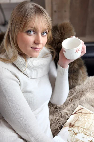 소파에 앉아 커피를 마시며 책을 읽고 있는 금발의 여자 — 스톡 사진
