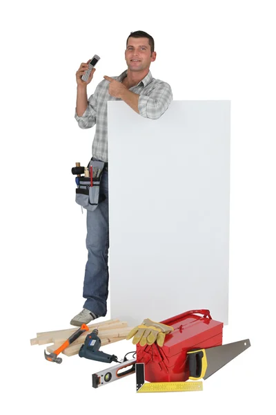 Carpenter pointing to mobile telephone whilst stood by blank poster — Stock Photo, Image