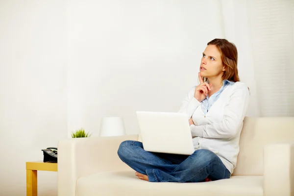 Молодая взрослая женщина думает на диване с ноутбуком — стоковое фото