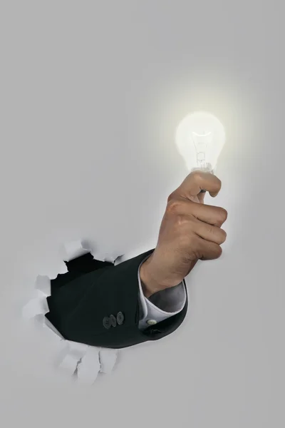 Empresário mão quebrando através de uma parede de papel segurando lâmpada — Fotografia de Stock