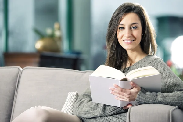 Portrait of a happy young woman lying on couch with book — Stock Photo, Image