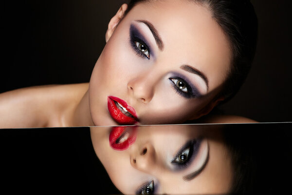 High fashion look.glamour fashion portrait of beautiful sexy brunette girl with bright makeup and red lips and her reflection in mirror table on dark