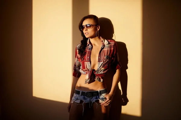 stock image Portrait of beautiful fashion sexy brunette girl in jeans and colorful shirt in glasses behind sunset indoors