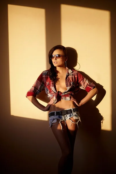 Retrato de moda bonita menina morena sexy em jeans e camisa colorida em óculos atrás do pôr do sol dentro de casa — Fotografia de Stock