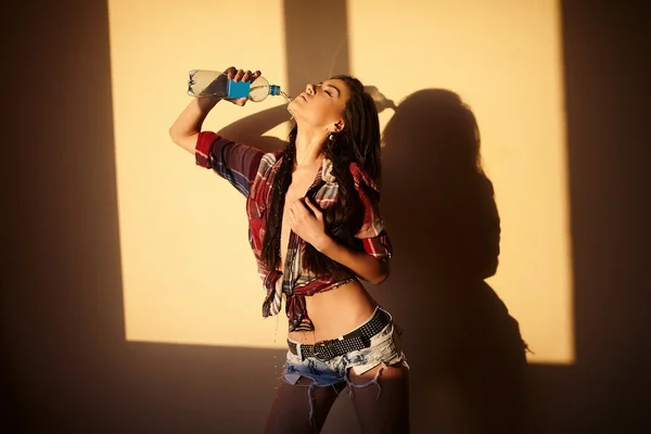 아름 다운 패션 섹시 한 갈색 머리 소녀 청바지와 병 물 실내로 일몰 뒤에 화려한 셔츠의 초상화 — 스톡 사진