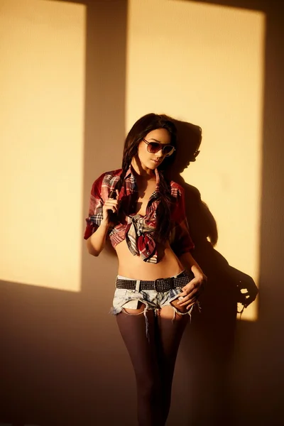 Portrét krásné módní sexy tmavovláska v džínách a barevné tričko v brýlích za západu slunce uvnitř — Stock fotografie
